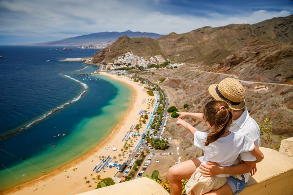 Mother and daughter sitting on a ledge, looking out over Las Teresitas beach in Tenerife, pointing towards the beach. Traveling with children concept